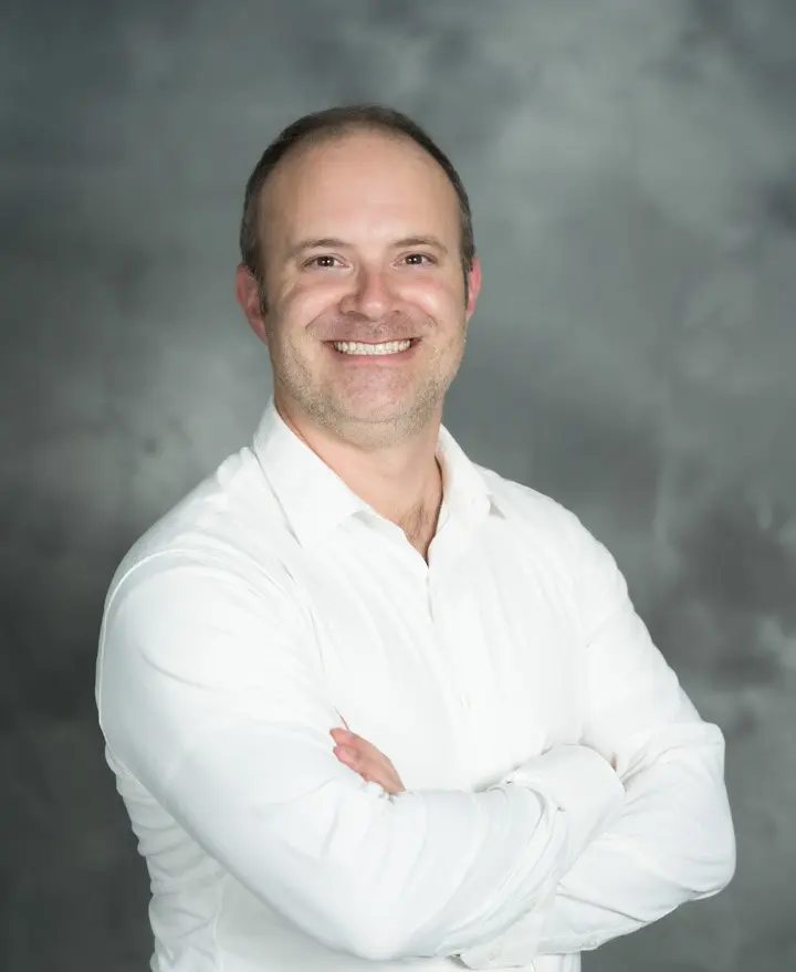 Smiling man in a white shirt with arms crossed against a gray background.